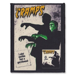 Op de afbeelding: Een zwart-witte patch met een bandlogo voor The Cramps. Het logo is geel en de tekst zegt "The Cramps". De patch toont een zwart-witte afbeelding van twee figuren met groene huid. De tekst "I ain't nothin' but a gorehound" staat op de patch.