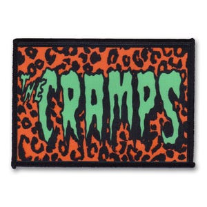 Könnte beinhalten: Ein schwarzer und orangefarbener Patch mit einem Leopardenmuster-Hintergrund. Das Wort "The Cramps" ist in grünen Buchstaben mit schwarzen Umrissen geschrieben.