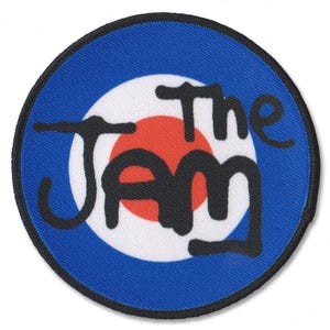 May include: A circular embroidered patch featuring a target design with a red centre, white ring, and blue outer border. The black text "The Jam" is written across the centre in a stylised, hand-drawn font, with a black stitched border edge.
