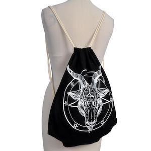 Może przedstawiać: Czarny worek ze sznurkiem z białą ilustracją głowy kozła w pentagramie. Torba ma białe sznurki i jest pokazana na manekinie. Akcesorium w stylu gotyckim, prawdopodobnie wykonane z tkaniny.