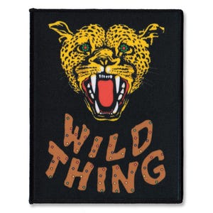 Peut inclure: Écusson rectangulaire noir avec un visage de léopard rugissant jaune et noir avec des yeux verts. Le texte "WILD THING" est en lettres marron avec de petits ornements ronds de couleur or.