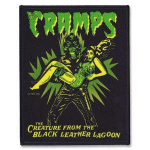 Puede incluir: Parche bordado negro y verde con un monstruo verde sosteniendo a una mujer con un vestido amarillo. El parche tiene el texto "The Cramps" y "The Creature From The Black Leather Lagoon" en verde.