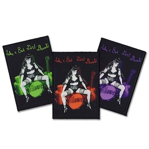 May include: Three black fabric patches with the text "Like a Bad Girl Should" in white cursive lettering. Each patch features a black and white illustration of a woman sitting on a drum with a guitar behind her. The drums are colored green, red, and purple.