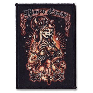 Pode incluir: Remendo retangular preto com uma ilustração no estilo Dia de los Muertos. A imagem retrata um esqueleto feminino com cabelo comprido, segurando um coração. O texto "Muerte Eterno" está no topo, com rosas e uma cruz na parte inferior.