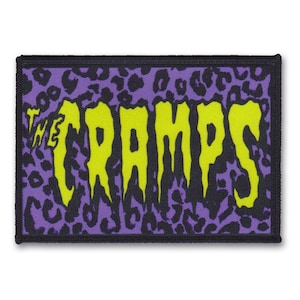 May include: A purple and black patch with a yellow "The Cramps" logo. The logo is in a dripping font and has a leopard print background.