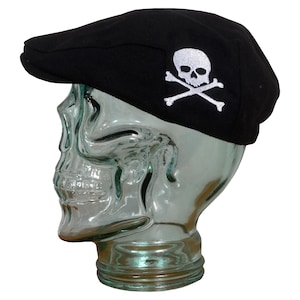 May include: A black newsboy cap with a white skull and crossbones embroidered on the side. The cap is displayed on a glass skull head. The hat has a curved brim and a rounded crown.