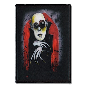 May include: A rectangular fabric patch featuring a stylised illustration of a pale, bald figure with large dark eyes and sharp fangs. The figure wears a black high-collared garment against a vibrant red and dark grey background with splatter effects.