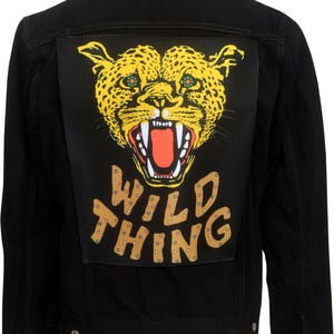 Pode incluir: Casaco de ganga preto com um grande patch nas costas com a cara de um leopardo amarelo com a boca aberta e as palavras "WILD THING" em letras castanhas.