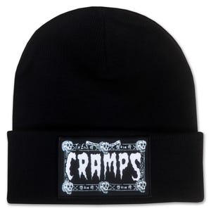 May include: Black knit beanie featuring a rectangular patch with the band name "CRAMPS" in a gothic font. The patch is framed with skulls and decorative elements, creating a punk rock aesthetic.