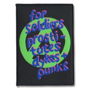 May include: A rectangular black fabric patch featuring a bright green circle with a black anarchy symbol inside. The text "for soldiers prosti-tutes dykes + punks" is printed in blue with pink outlines, layered over the green circle design.