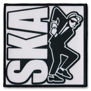 Op de afbeelding: Vierkante patch met het woord "SKA" in grote witte blokletters. Een zwart-wit silhouet van een man in pak, hoed en zonnebril wordt afgebeeld in een wandelhouding. De patch heeft een zwarte rand.