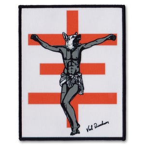 以下が含まれることがあります： 赤い十字架と、十字架の上で犬の頭をした人物の白黒のイラストが描かれた白いパッチ。右下に「Val Demarco」の文字があります。