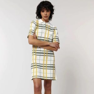 May include: A white polo dress with a yellow and gray plaid pattern. The dress has short sleeves and a collared neckline. The model is standing with arms crossed, showcasing the dress's design and fit.