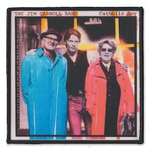 May include: A rectangular embroidered patch featuring the album cover for The Jim Carroll Band's Catholic Boy. The image shows three people standing together, with the person on the left in a bright blue coat and the person on the right in a red coat.