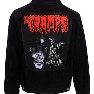 Puede incluir: Chaqueta vaquera negra con un parche grande en la espalda con el nombre de la banda "The Cramps" en rojo, con letras en forma de llama. Debajo, un gráfico en blanco y negro con el texto "You Ain't No Punk You Punk".