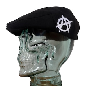 Könnte beinhalten: Schwarze Newsboy-Cap mit einem weißen, aufgestickten Anarchie-Symbol auf der Seite. Die Cap hat eine runde Krone und einen kurzen, steifen Schirm. Die Mütze wird auf einem Glaskopf in Schädelform präsentiert.