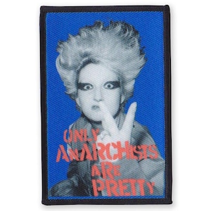 May include: A blue and black embroidered patch featuring a black and white image of a person with a mohawk hairstyle. The person is making a peace sign with their hand. The text "Only Anarchists Are Pretty" is printed in red and pink on the patch.