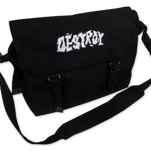 Pode incluir: Bolsa mensageiro preta com a palavra "DESTROY" bordada em branco na aba. A bolsa tem uma alça de ombro longa e ajustável e dois fechos de fivela. A bolsa é feita de tecido durável.