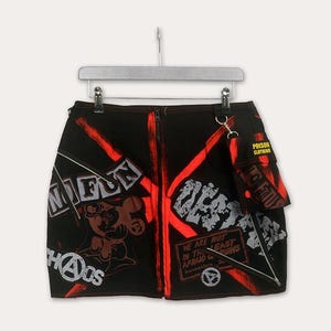 Puede incluir: Minifalda negra con diseños gráficos rojos y blancos. La falda tiene una cremallera frontal, un pequeño bolsillo y varios elementos de texto, incluyendo "FUN" y "CHAOS". El diseño tiene una estética punk rock.