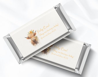 May include: A white chocolate bar wrapper with a watercolor illustration of a Highland cow with flowers in its hair. The text "Holy Cow! They are having a baby! Janet and Dave 2023" is printed in gold on the wrapper.