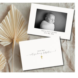 May include: A white card with a black and white photo of a baby. The text on the card reads "Emelia Grace Rylands 27th May 2024" and "Uncle Matt will you be my Godfather?" with a gold cross.