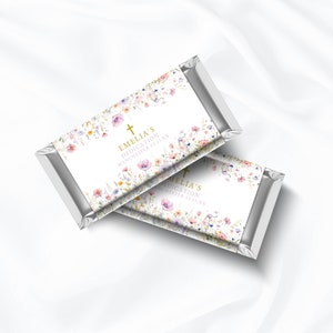 Puede incluir: Dos barras de chocolate envueltas en papel plateado con etiquetas blancas con un diseño floral de acuarela y el texto "Emelia's Dedication Manchester 11.11.xx" y una cruz dorada.