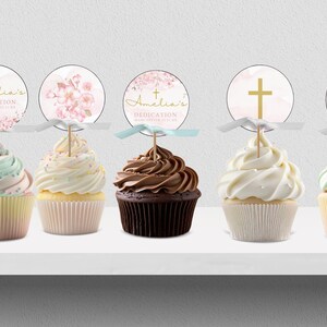 May include: Five cupcakes decorated with pink and white frosting and topped with round, pink and white paper cupcake toppers with gold lettering that reads "Amelia's Dedication" and a gold cross.