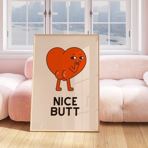 Puede incluir: Una impresión enmarcada con un personaje de corazón de dibujos animados con un cuerpo rojo y una expresión de sorpresa. El personaje está de pie con las manos cubriéndose la boca. El texto "NICE BUTT" está impreso debajo del personaje.
