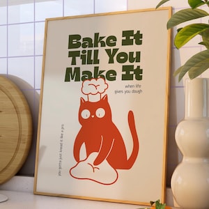 May include: A framed poster featuring the text "Bake It Till You Make It" and a cartoon cat wearing a chef's hat, kneading dough. The poster has a cream background and a wooden frame. The text "when life gives you dough" is also included.