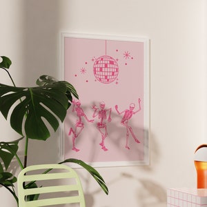 Puede incluir: Una impresi&oacute;n enmarcada en rosa con una bola de discoteca y tres esqueletos bailando. La impresi&oacute;n est&aacute; colgada en una pared blanca. Una planta verde est&aacute; en primer plano. Una silla verde claro est&aacute; en primer plano. Una peque&ntilde;a l&aacute;mpara naranja est&aacute; sobre una superficie de azulejos blancos y rosas.