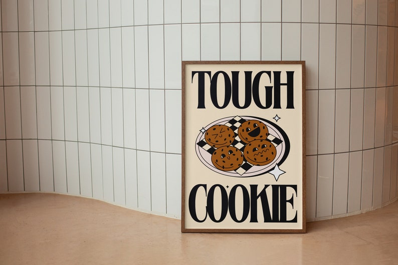 May include: A framed print with the text "TOUGH COOKIE" in black bold letters. The print features a cartoon illustration of four chocolate chip cookies with faces on a pink plate with a black and white checkered pattern.