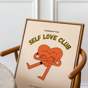 May include: A framed print with a light brown background featuring a cartoon heart hugging itself. The text "MEMBERSHIP PASS SELF LOVE CLUB" is in a dark green font. The text "I am worthy of all things good" is in black font at the bottom of the print.