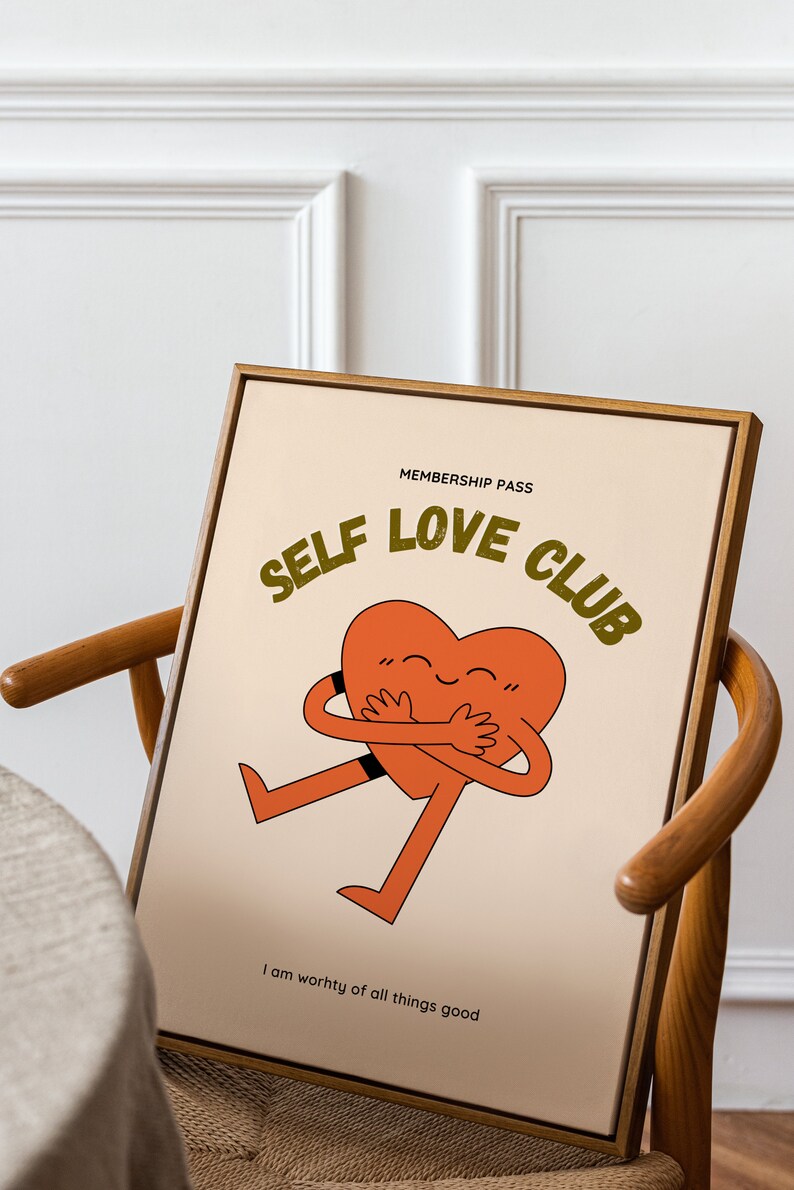 May include: A framed print with a light brown background featuring a cartoon heart hugging itself. The text "MEMBERSHIP PASS SELF LOVE CLUB" is in a dark green font. The text "I am worthy of all things good" is in black font at the bottom of the print.