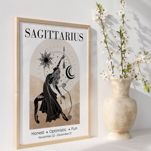 May include: A framed Sagittarius zodiac art print. The print features a black illustration of a figure with a bow and arrow, set against a beige and brown background. The word "SAGITTARIUS" is at the top, with "Honest + Optimistic + Fun" and the dates November 22 - December 21 below.