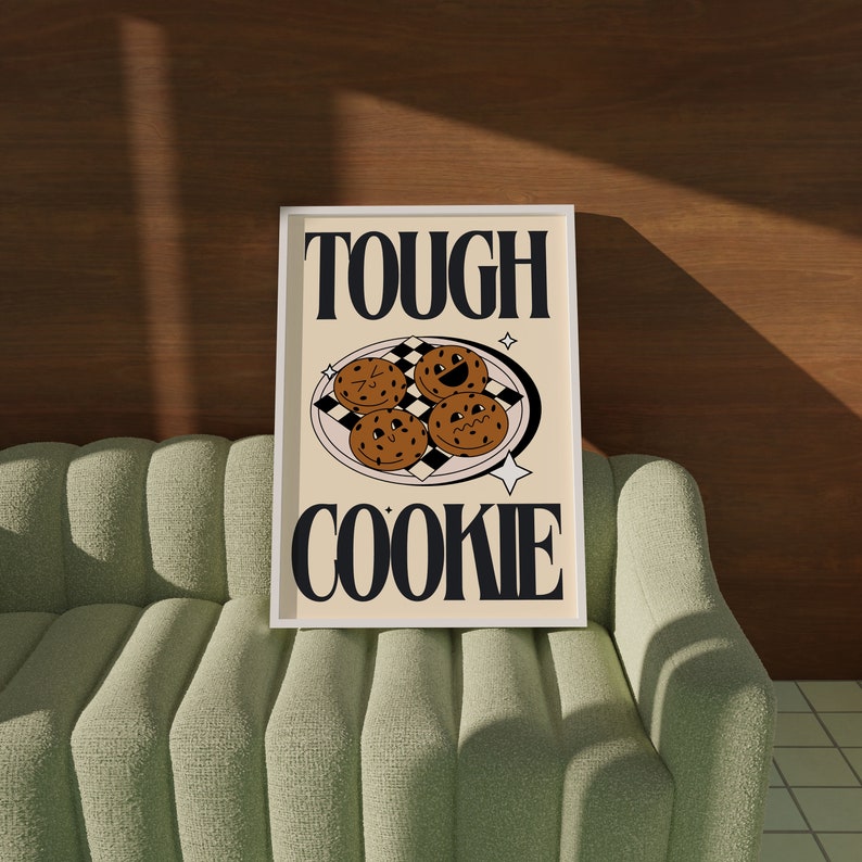 May include: A white framed print with the text "Tough Cookie" in black letters. The print features a black and white checkered plate with four chocolate chip cookies with faces on them.