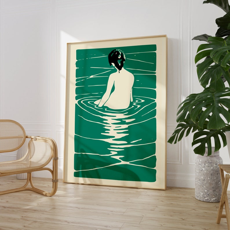 May include: A green and white abstract print of a woman sitting in a pool of water. The woman is facing away from the viewer and has dark hair. The print is framed in a light wood frame.