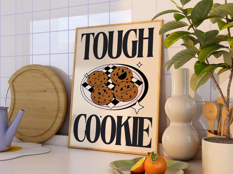 May include: A beige poster with black text that reads "TOUGH COOKIE" and a graphic of four chocolate chip cookies with faces on a checkered plate. The poster is framed in a light brown wood frame.