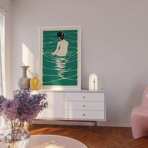 May include: A white dresser with three drawers and gold handles. A white lamp with a gold arch sits on top of the dresser. A framed print of a woman in a pool of water hangs on the wall above the dresser. The print has a green background.