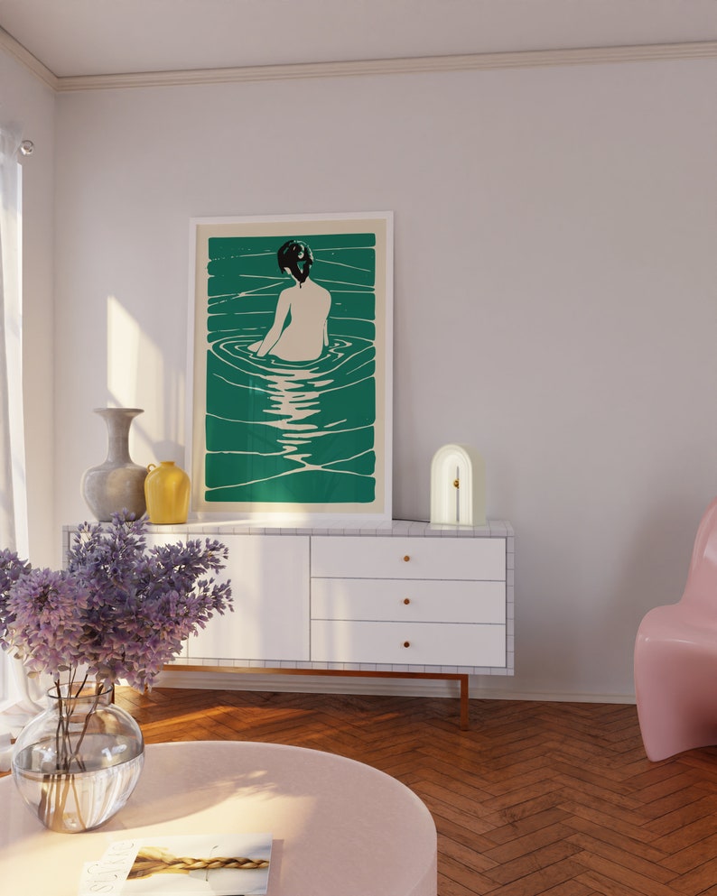 May include: A white dresser with three drawers and gold handles. A white lamp with a gold arch sits on top of the dresser. A framed print of a woman in a pool of water hangs on the wall above the dresser. The print has a green background.