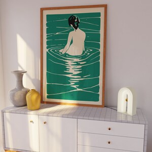 May include: A framed print of a woman sitting in a pool of water. The woman is facing away from the viewer and has dark hair. The water is a vibrant green color and the background is a light cream color.