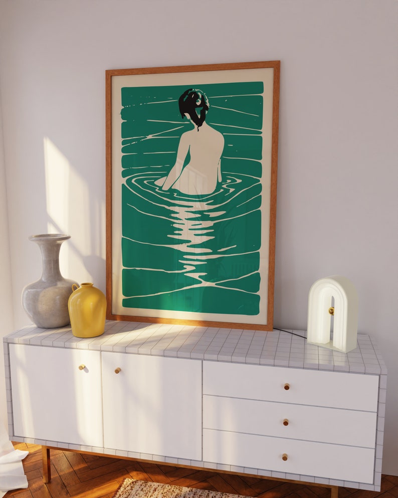 May include: A framed print of a woman sitting in a pool of water. The woman is facing away from the viewer and has dark hair. The water is a vibrant green color and the background is a light cream color.