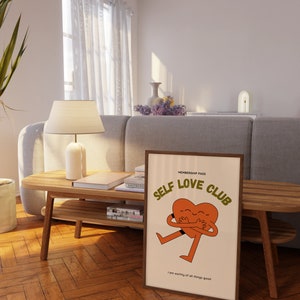 May include: A framed print with a beige background featuring a cartoon heart character hugging itself. The text "MEMBERSHIP PASS SELF LOVE CLUB" is in orange and brown text. The text "I am worthy of all things good" is in smaller brown text at the bottom of the print.