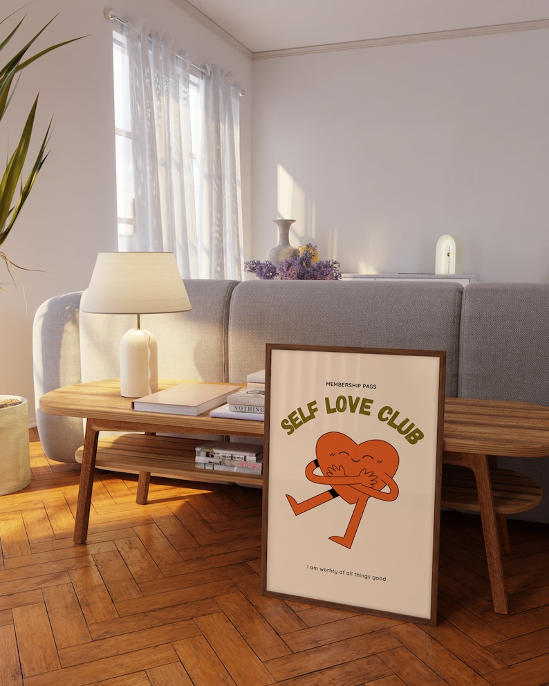 May include: A framed print with a beige background featuring a cartoon heart character hugging itself. The text "MEMBERSHIP PASS SELF LOVE CLUB" is in orange and brown text. The text "I am worthy of all things good" is in smaller brown text at the bottom of the print.