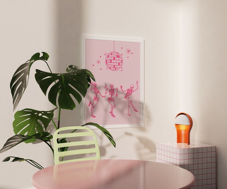 Puede incluir: Una impresi&oacute;n enmarcada en rosa con una bola de discoteca y tres esqueletos bailando. La impresi&oacute;n est&aacute; colgada en una pared blanca. Una planta verde est&aacute; en primer plano. Una silla verde claro est&aacute; en primer plano. Una peque&ntilde;a l&aacute;mpara naranja est&aacute; sobre una superficie de azulejos blancos y rosas.