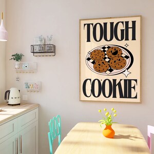 May include: A framed print with the text "TOUGH COOKIE" in black letters on a beige background. The print features a plate of four chocolate chip cookies with smiling faces.