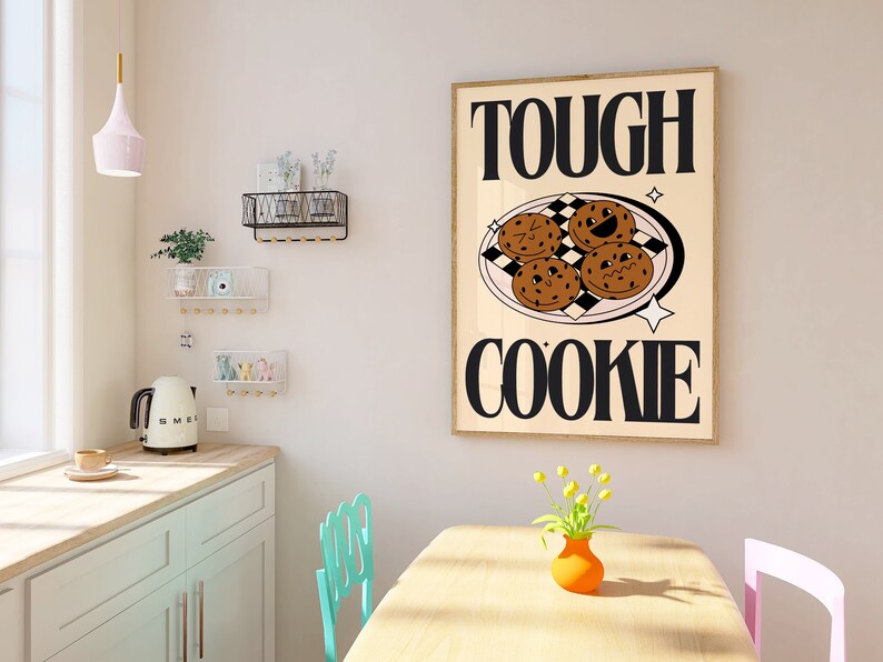 May include: A framed print with the text "TOUGH COOKIE" in black letters on a beige background. The print features a plate of four chocolate chip cookies with smiling faces.