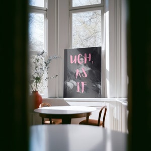 Puede incluir: Un p&oacute;ster en blanco y negro con las palabras "ugh, as if" en letras rosas. El p&oacute;ster est&aacute; colgado en una pared blanca en una habitaci&oacute;n con una mesa y sillas.