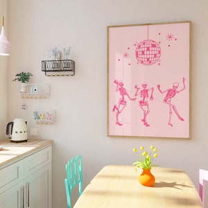 Puede incluir: Una impresi&oacute;n enmarcada en rosa con una bola de discoteca y tres esqueletos bailando. La impresi&oacute;n est&aacute; colgada en una pared rosa claro en una cocina con una mesa y sillas de madera clara.