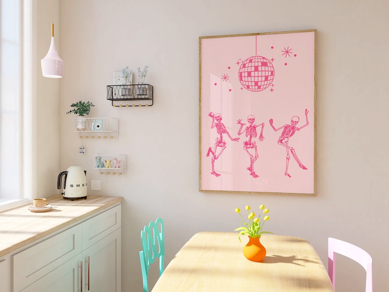 Puede incluir: Una impresi&oacute;n enmarcada en rosa con una bola de discoteca y tres esqueletos bailando. La impresi&oacute;n est&aacute; colgada en una pared rosa claro en una cocina con una mesa y sillas de madera clara.