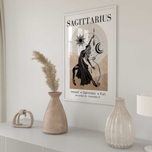 May include: A framed Sagittarius zodiac art print. The black and white illustration depicts a figure with a bow and arrow, set against a beige and brown background. The print includes the word "Sagittarius" and the traits "Honest, Optimistic, Fun."